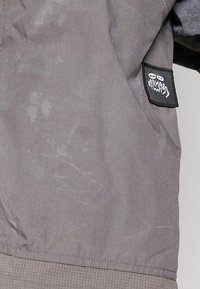 Gray jacket featuring a textured finish, visible creases, and a black logo patch on the sleeve. Fabric has a slightly glossy appearance.