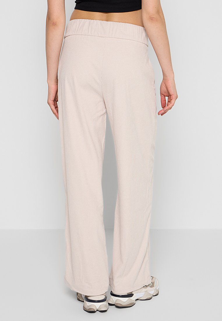 Donna che indossa pantaloni beige a gamba larga e sneakers voluminose, in piedi con la schiena rivolta verso la telecamera su uno sfondo semplice.