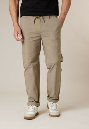 Homme portant un pantalon beige à cordon de serrage, une chemise noire, des baskets blanches, debout avec une main dans la poche devant un fond uni.