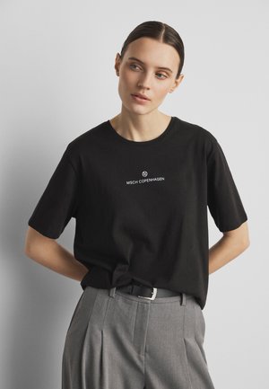 Ung kvinde med mørkt hår iført en sort MSCH Copenhagen t-shirt, stoppet ned i grå plisserede bukser med sort bælte, stående mod en ensfarvet baggrund.