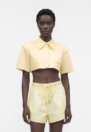 Kvinde iført lysegul cropped kortærmet skjorte og matchende højtaljede shorts står mod hvid baggrund.