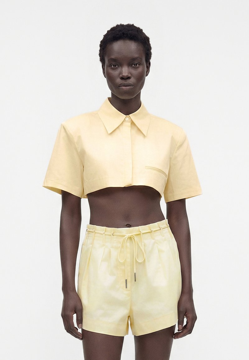 Femme portant un t-shirt court à manches courtes jaune pâle et un short taille haute assorti, debout sur fond blanc.