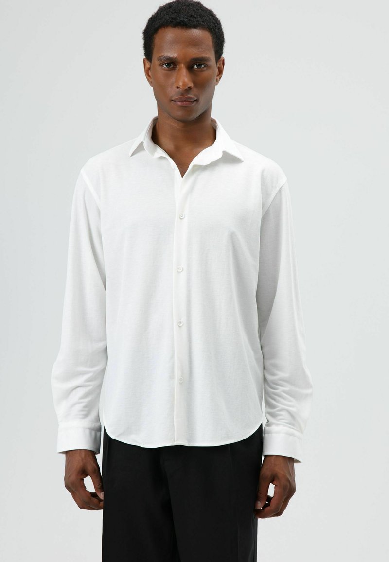 Witte blouse met lange mouwen van lichtgewicht stof, voorzien van een knoopsluiting aan de voorkant, afgeronde zoom en een klassieke kraag. Draagt zwart broek.