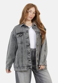 Veste en denim gris avec de grandes poches avant, des boutons et une coupe décontractée. Associée à un haut blanc et un jean gris assorti.