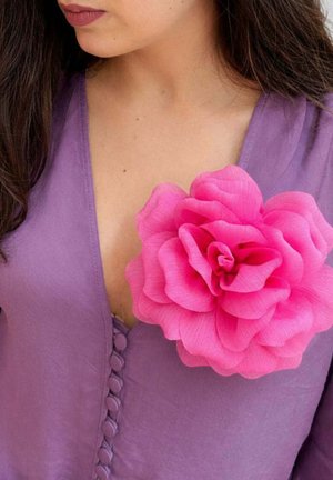 Kobieta ubrana w fioletowy top z dekoltem w serek, ozdobiony guzikami pokrytymi materiałem, z dużym, jasnoróżowym kwiatem z tkaniny przypiętym blisko dekoltu.
