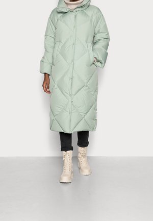 Frau trägt hellgrüne gesteppten langen Steppmantel, schwarze hochgekrempelte Hose, beige Schnürstiefel und cremefarbenen Rollkragenpullover.