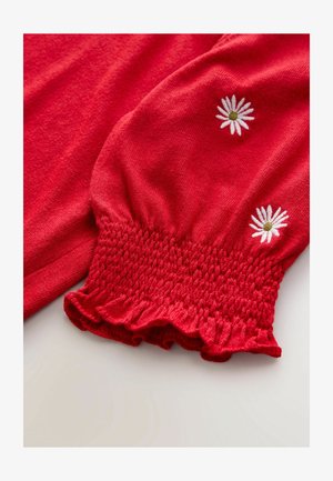 Red long-sleeve garment featuring smocked detailing at the wrist and floral embroidery with white daisy designs on the sleeves.