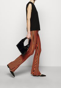 Svart ärmlös topp, orange och svart randiga vida byxor, och en glansig svart handväska med en tvistad handtag. Slim-fit siluett.