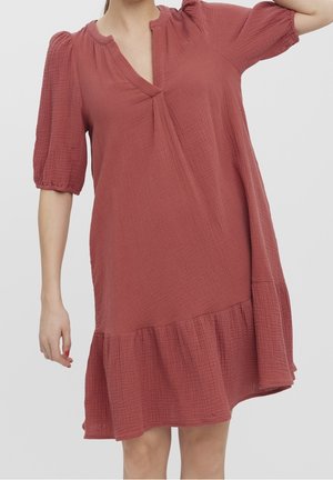 Femme portant une robe courte à manches courtes, texturée et ample, de couleur rose poussiéreux, avec un décolleté en V et un ourlet à volants, sur fond uni.