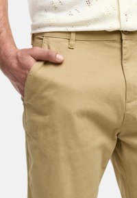 Khaki chinos z gładkiej, bawełnianej tkaniny. Posiadają przednie kieszenie, płaski przód i szlufki na pasek. Zdjęcie zbliżeniowe dłoni opierającej się na kieszeni.