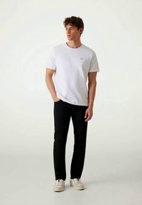 Homem em pé, vestindo uma t-shirt branca de manga curta simples, calças pretas e ténis brancos, olhando para o lado com uma mão no bolso.