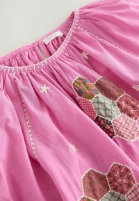 Blouse en coton rose avec col froncé, présentant un patchwork hexagonal de motifs floraux et à carreaux. Accents de couture blanche partout.