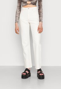 Jeans brancos de cintura alta com um design de perna reta, feitos de denim. O modelo está a usar sandálias de plataforma pretas com tiras largas.