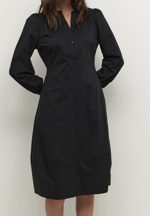 Frau trägt ein schwarzes, langärmliges, knielanges Kleid mit aufgepufften Schultern und einem kurzen, geknöpften Schlitz am Ausschnitt.