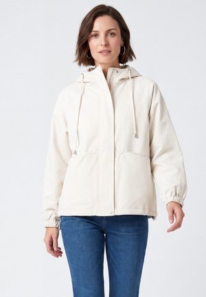 Donna con capelli corti castani che indossa una giacca con cappuccio beige chiaro e jeans blu, in piedi davanti a uno sfondo bianco semplice.