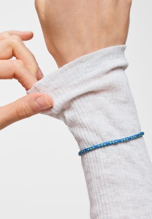 Ręka ściąga biały prążkowany rękaw na nadgarstek, na którym noszona jest niebiesko-zielona wzorzysta bransoletka na jasnej skórze.