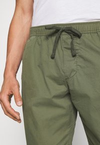 Close-up van een persoon die olijfgroene drawstring broek en een wit shirt draagt, met hun linkerhand ontspannen naast zich.
