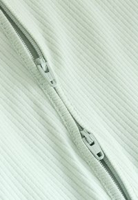 Ljusgrön zip-up-plagg med ribbad textur. Har plastdragkedja och toppsöm längs kanterna vilket ger en strukturerad look.