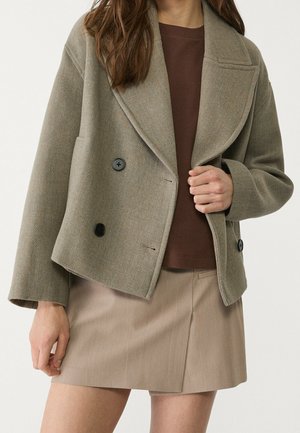 Kobieta w taupe dwurzędowej wełnianej kurtce na brązową koszulę i beżową mini spódniczkę w stylu kopertowym.