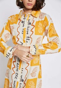 Long-sleeve shirt in cream fabric featuring yellow and orange abstract floral designs, buttoned with a collared neck and pleated cuffs.