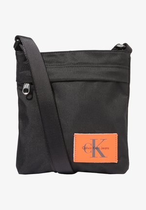 Sac bandoulière en tissu noir avec fermeture éclair, sangle réglable, et patch logo orange Calvin Klein Jeans à l'avant.