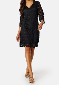 Robe en dentelle noire avec décolleté en V, manches trois-quarts, ourlet festonné et motif floral. Le modèle porte des sandales minimalistes à brides.