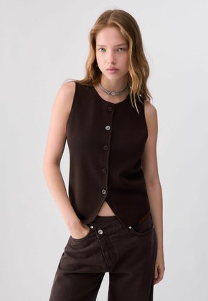 Giovane donna con lunghi capelli ondulati indossa un gilet senza maniche marrone scuro con bottoni e pantaloni coordinati, in piedi con una mano in tasca.
