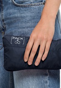 Mörkblå, lätt och packbar påse med en logotyp, med en slät struktur och dragsko, hålls i en hand mot denim.