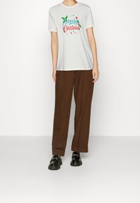 Bílé tričko s nápisem "Veselé Vánoce" v barevném písmu a designem cesmíny; spárováno s hnědými volnými kalhotami a černými platformovými botami.