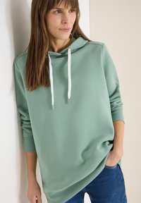 Jeune femme portant un sweat à capuche vert sauge avec des cordons blancs et un jean bleu, debout contre un mur de couleur claire, mains dans les poches.