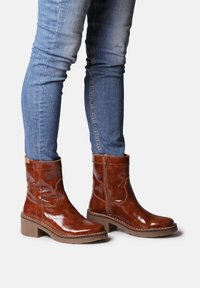 Brown leather ankle boots with a glossy finish, side zipper, and a chunky sole, worn with blue skinny jeans.