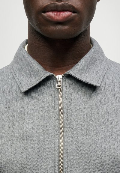 Tuvumā redzam personas apakšējo sejas daļu un kaklu, kas vilkusi pelēku zipa jaku ar strukturētu audumu un salocītu apkakli.