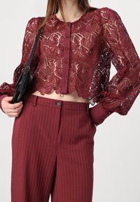 Blusa corta de encaje burdeos con estampado floral, cierre de botones y mangas abullonadas. Combinada con pantalones a rayas a juego.