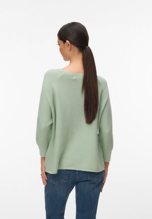 Femme aux longs cheveux foncés attachés en queue de cheval, portant un pull côtelé vert clair et un jean bleu, vue de dos sur fond blanc.