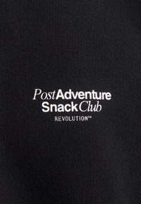 Tejido negro con textura acanalada que presenta un texto bordado en blanco: "Club de Bocadillos Post Aventura REVOLUCIÓN™." Diseño sencillo y moderno.