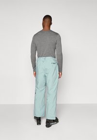 Pantaloni de schi de culoare albastru deschis, cu o croială relaxată, talie ajustabilă și accente cu logo, purtați împreună cu cizme de schi negre și o bluză gri cu mâneci lungi.