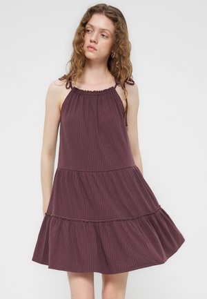 Mladá žena s vlnitými vlasmi nosiť bezrukávové, voľné fialové šaty s viazanými ramennými popruhmi a nariasenou viacvrstvovou sukňou.