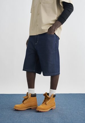 Uomo con pantaloncini blu scuro, camicia beige a maniche corte sopra una maglia nera a maniche lunghe, calzini bianchi e stivali da lavoro beige con lacci.