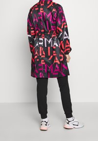 Osoba v černém kabátě s velkými červenými a fialovými písmeny, černých kalhotách a bílých teniskách, stojí na jednoduchém šedém pozadí.