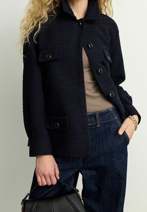 Marineblaue strukturierte Jacke mit spitzem Kragen, Frontknöpfen, zwei aufgesetzten Taschen, kombiniert mit einem beigen Oberteil und dunklen Jeans. Schwarze Tasche in der Hand.