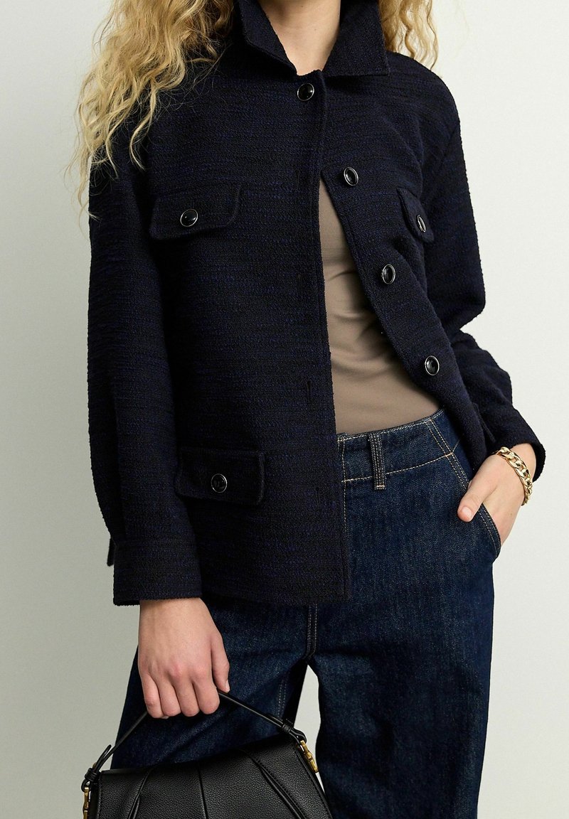 Mørkeblå tekstureret jakke med spids krave, frontknapper, to påsatte lommer, parret med en beige top og mørke jeans. Sort taske i hånden.