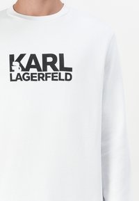 Sudadera blanca de manga larga con un atrevido estampado negro de "KARL LAGERFELD" junto a un perfil en silueta. Textura suave, cuello redondo.