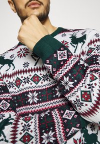 Strickpullover mit einem weißen Grundton, grünen Akzenten und einem festlichen Muster aus Schneeflocken und Rentieren in Rot, Grün und Schwarz.