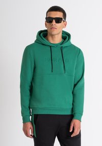 Sudadera con capucha verde de textura suave, con un bolsillo frontal, que presenta un logo en relieve tonal y cordones negros. Combinada con pantalones negros.
