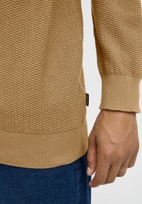 Pull en beige texturé avec une surface à motifs, des poignets et un ourlet côtelés. Le vêtement est associé à un jean bleu foncé.