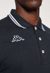 Polo nero con colletto a righe bianche, dotato di un piccolo logo ricamato. Ha bottoni beige e un tessuto testurizzato.