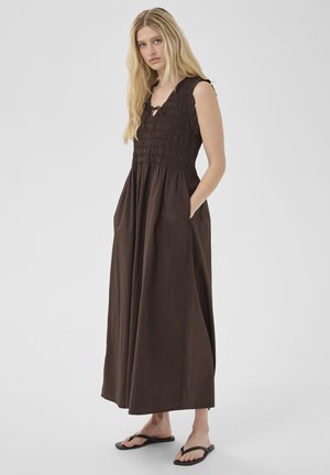 Kobieta o długich blond włosach, ubrana w bezrękawową, ciemnobrązową sukienkę maxi z marszczonym górnym stanem i kieszeniami, w połączeniu z czarnymi klapkami.