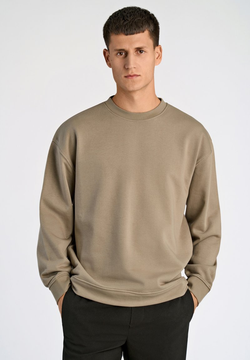 Felpa beige con scollo rotondo e maniche lunghe. Tessuto morbido e strutturato; vestibilità rilassata; indossata sopra pantaloni scuri. Design minimalista.