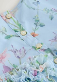 Svetlomodrá kvetinová látka s malými zlatými gombíkmi. Má volánový okraj a jemné kvetinové vzory v ružových, zelených a levanduľových odtieňoch.