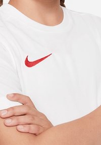 Bílý bavlněný tričko s červeným vyšívaným logem Nike na levé části hrudi, s klasickým designem kulatého výstřihu a krátkými rukávy.